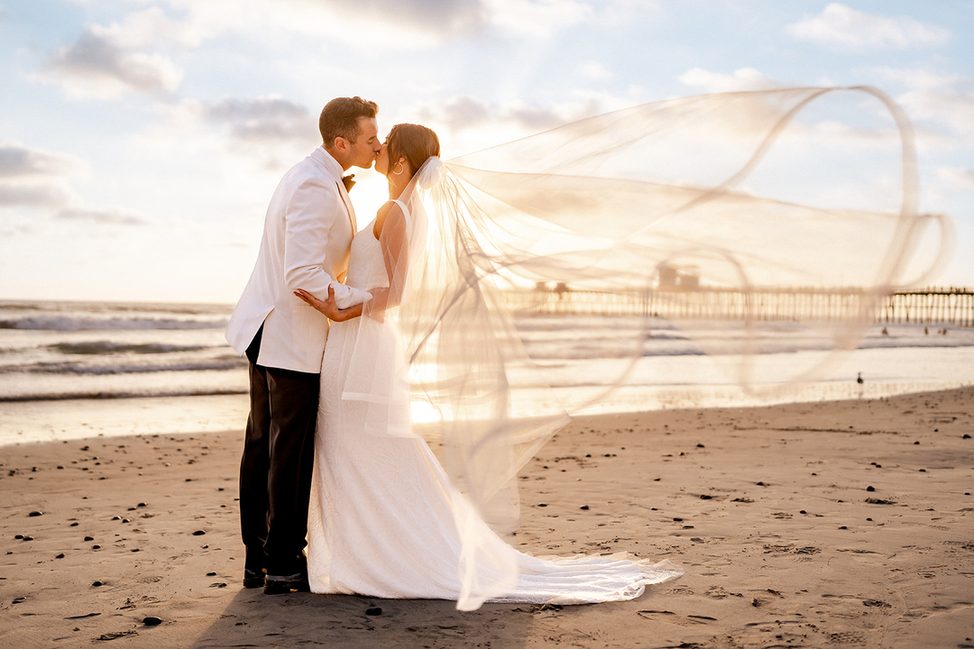 a wedding couple kissing by a beach while the bridal veil is blowing in the wind