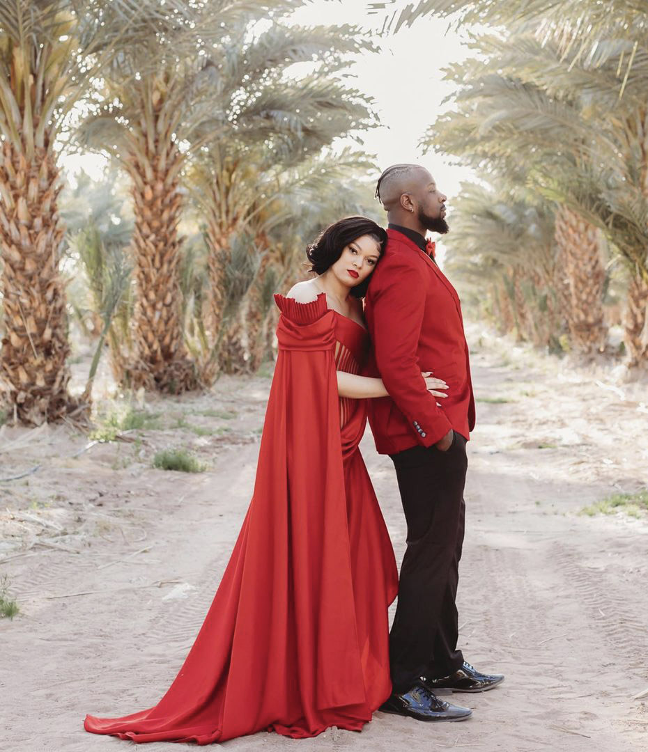 a couple standing in the middle of a row of trees in bright red wedding outfit