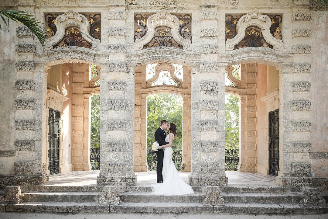 a newlywed couple kissing standing at the staircase of a beautiful stone structure in their wedding attire