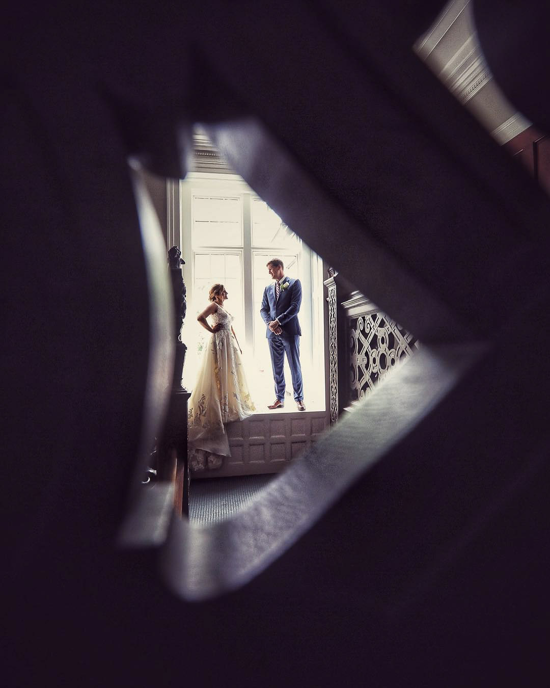 a wedding couple standing in front of a window seen through an creative opening in the wall