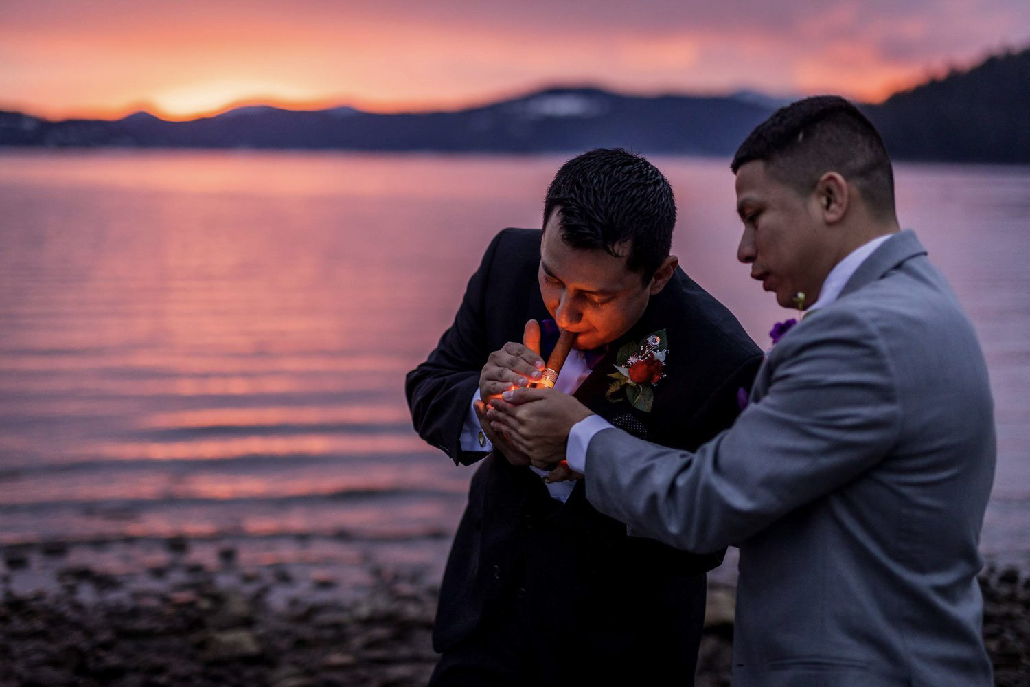 the best man helping the groom light a cigar beside a beautiful lake during the sunset