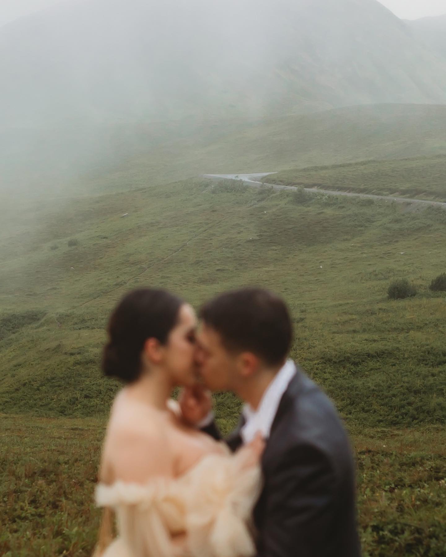a wedding couple kissing off focus in the foreground with foggy hilly landscape in the background