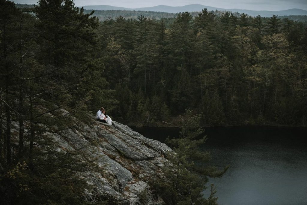 a couple sitting on a rocky cliff overlooking a lake surrounded by forest