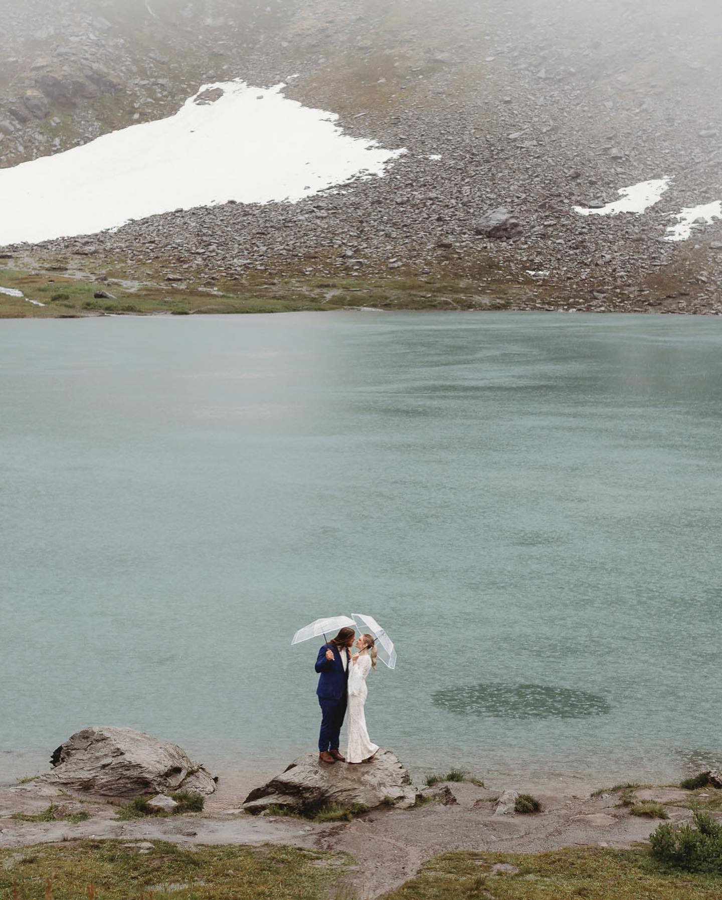 a wedding couple standing holding umbrellas in their wedding attire by a lake in rain
