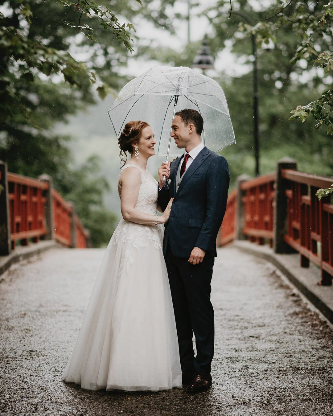 a wedding couple standing on a small bridge in their wedding attire sharing an umbrella