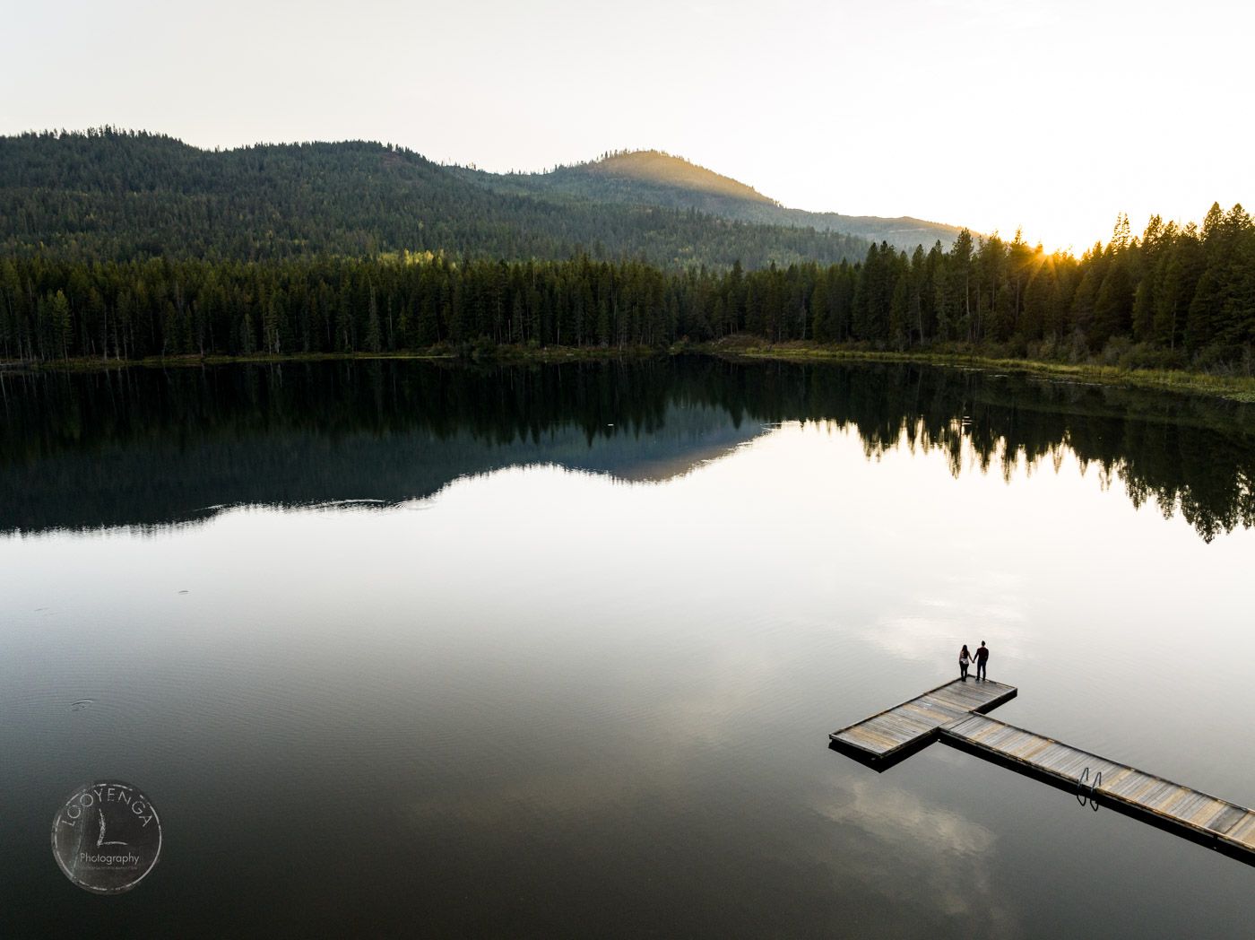 an aerial shot of a couple standing on a wooden pier on a lake surrounded by forest