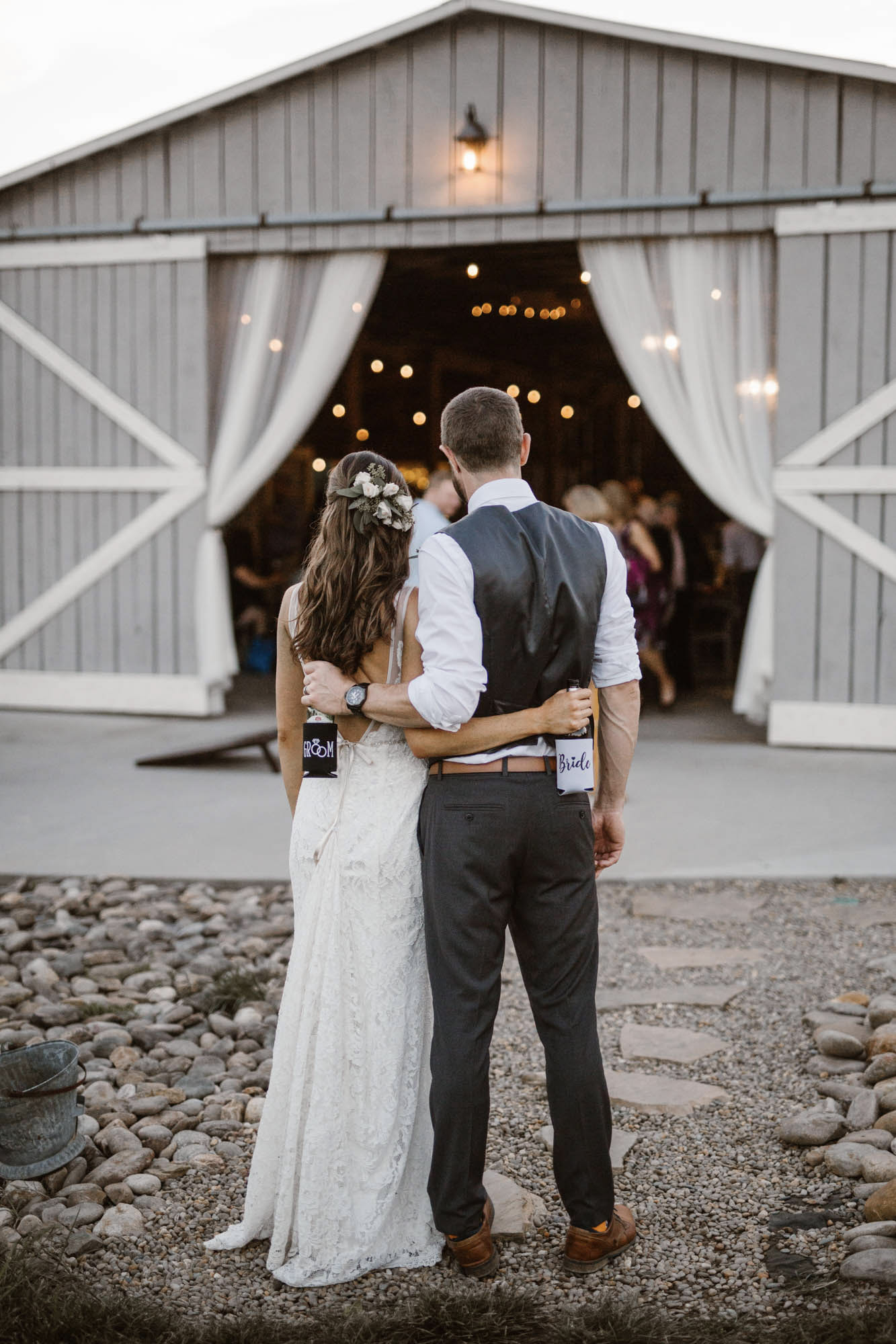 a wedding couple standing facing a barn holding a bride and a groom labeled bottles