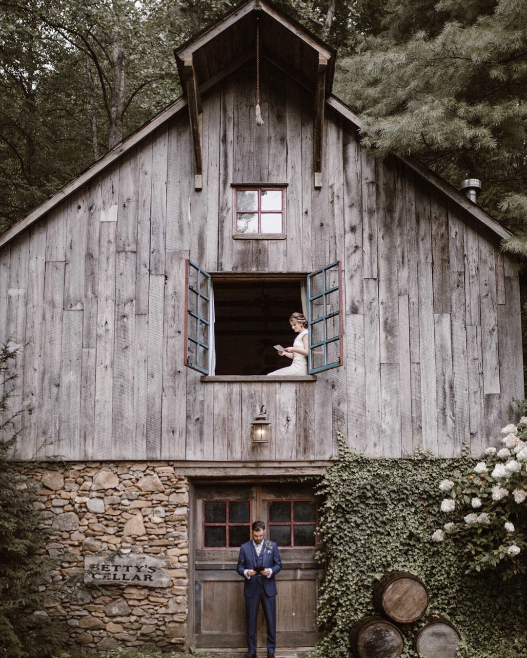a groom standing in front of a barn while the bride is sitting on the top window