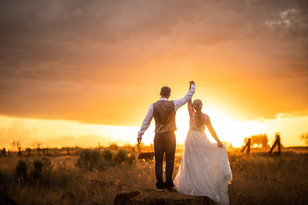 a wedding couple in their wedding attire raising their hands together facing the sunrise
