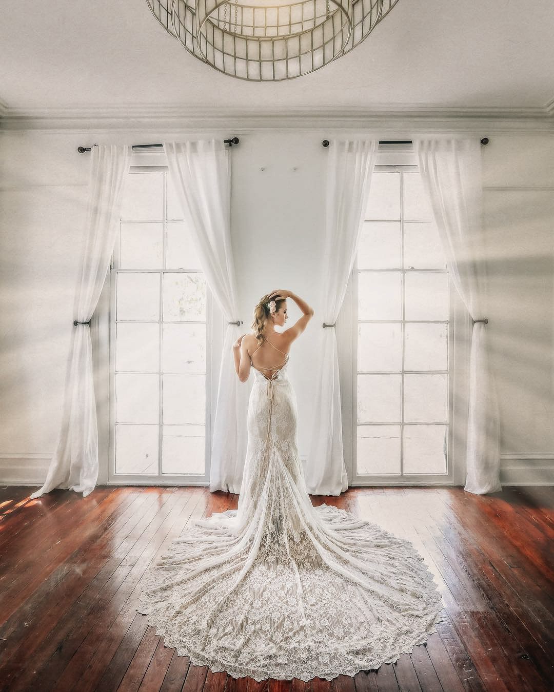 a bride posing with the train of her wedding dress spread out while light shines from two windows behind her