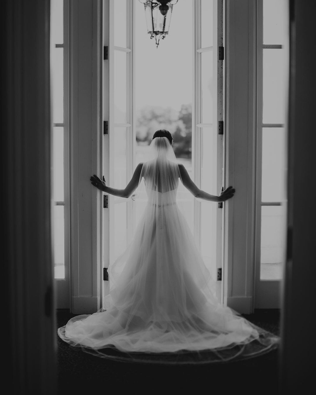 a black and white image of a bride opening the front door in her wedding dress