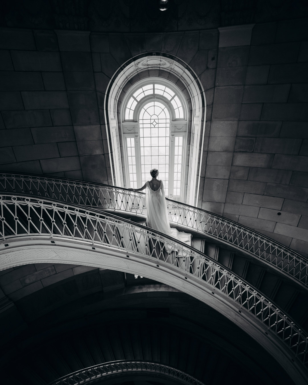 a bride standing on a spiral stair case looking outside through a giant arched window